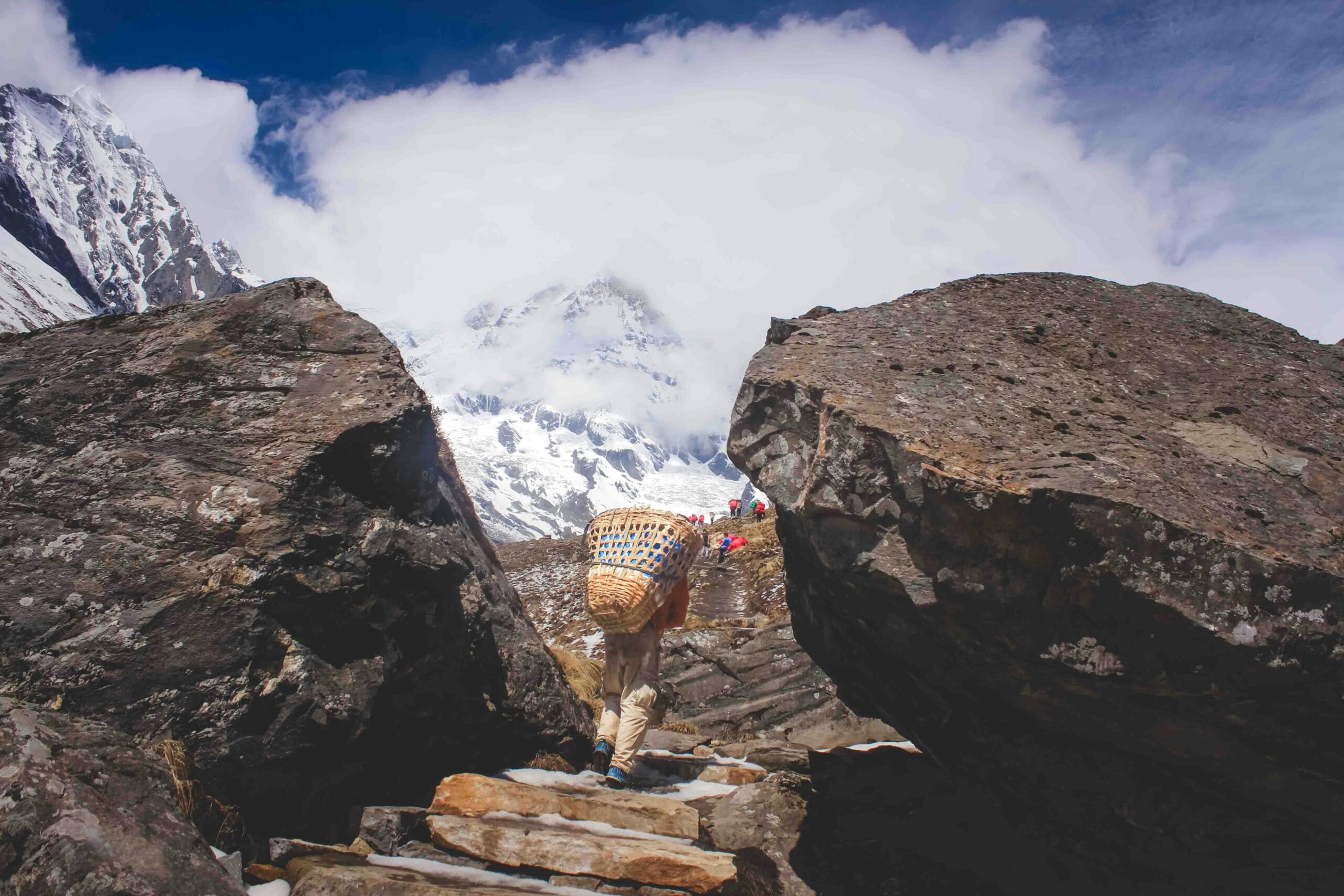 Trekker carrying basket climbs rocky path in Kathmandu's mountainous terrain.