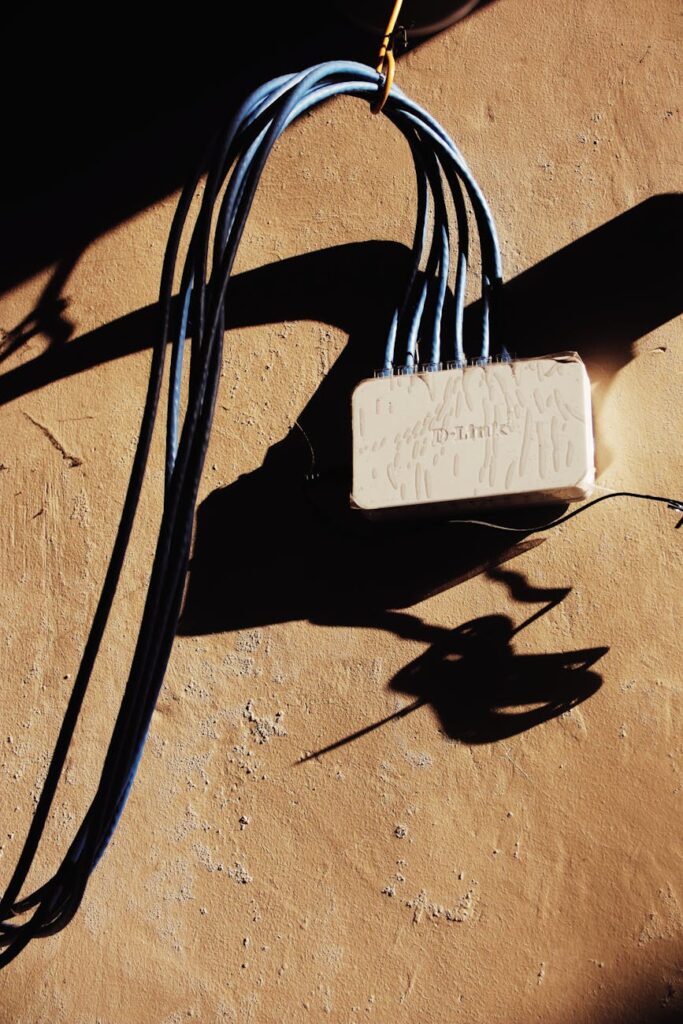 White electrical network adapter with various blue wires hanging on rough brown wall of building with bright sunlight and shadow